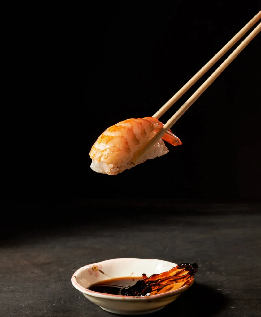 Prawn nigiri held with chopsticks above a dish of soy sauce