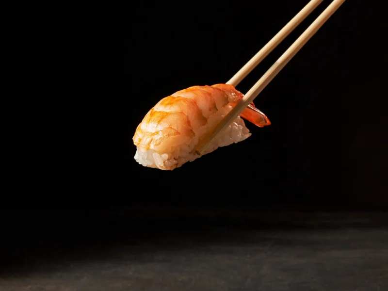 Prawn nigiri held with chopsticks above a dish of soy sauce